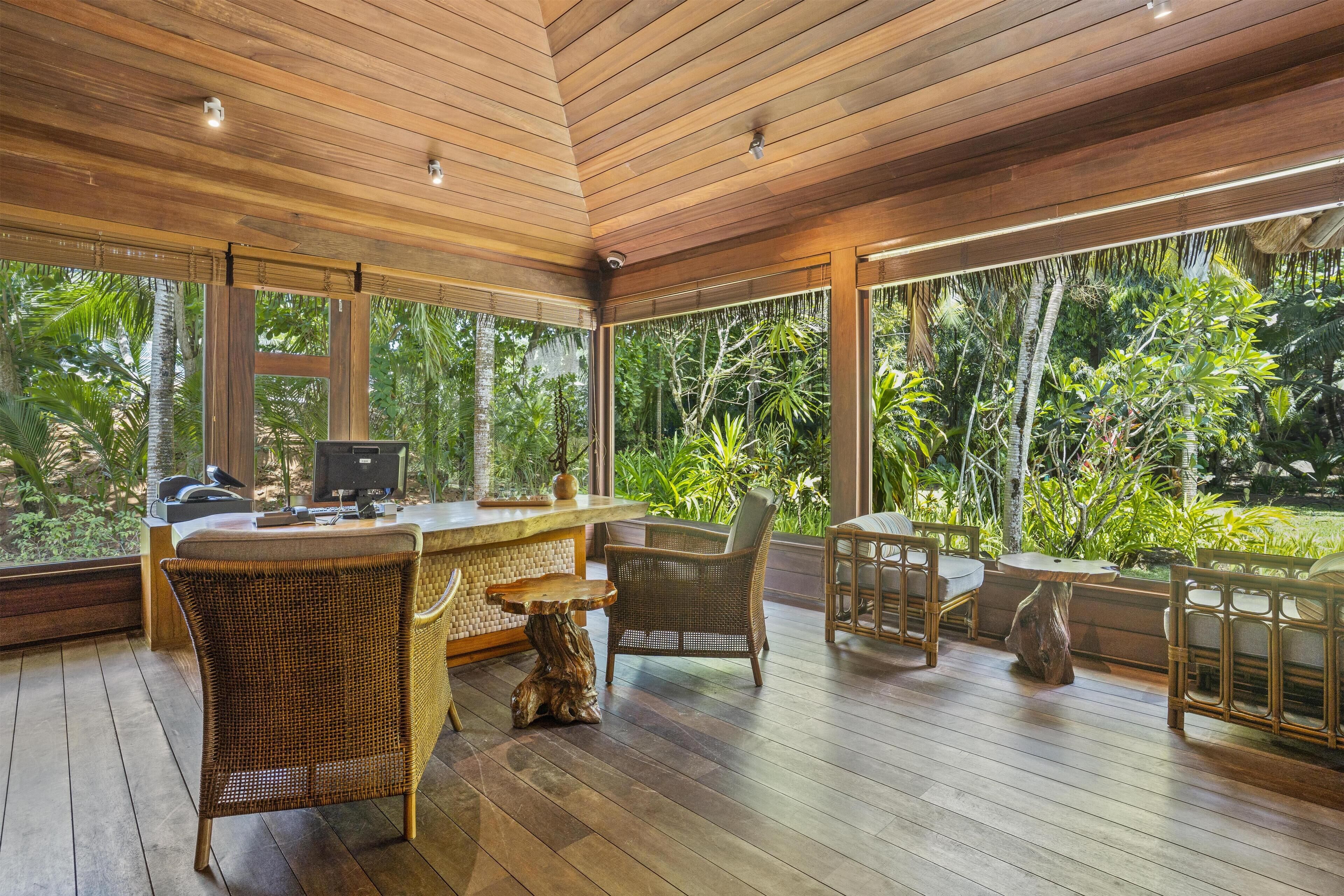 A wooden-paneled room with a bar area, wicker furniture, and large windows overlooking a lush garden.