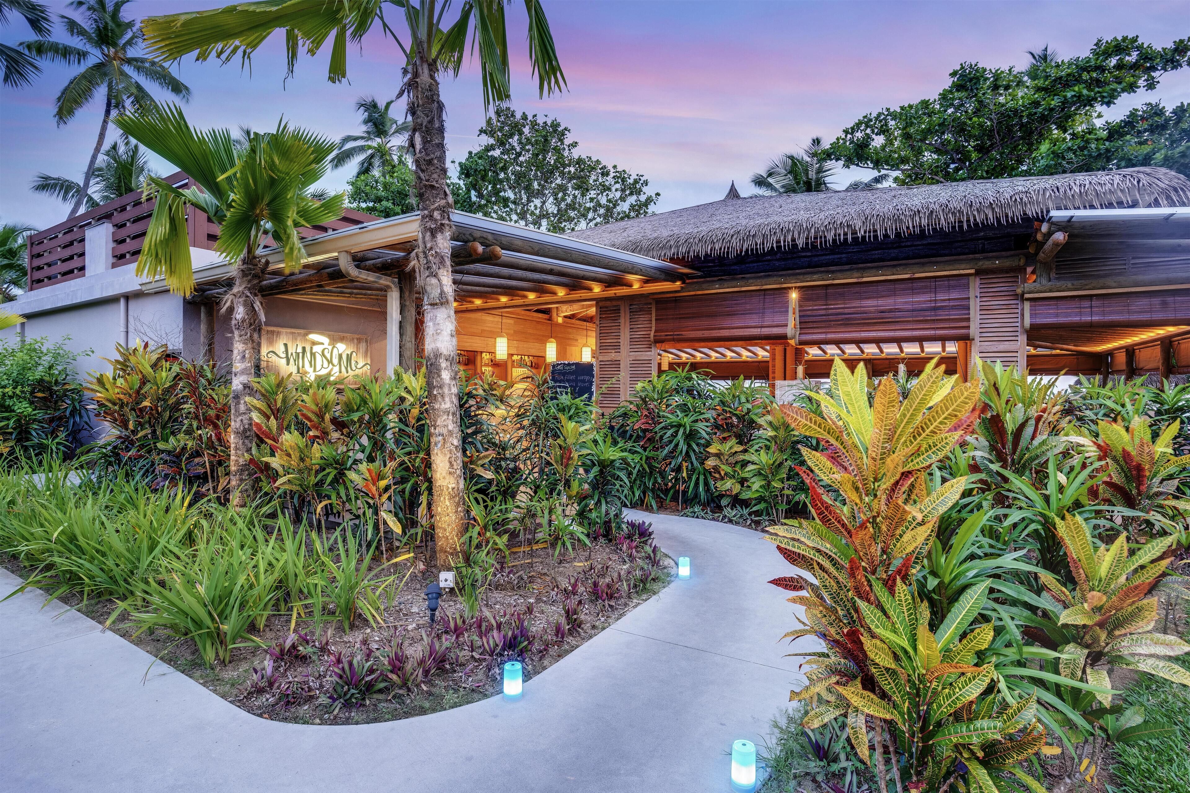 Un resort con una estructura de techo de paja, rodeado de exuberante vegetación tropical y un sendero pavimentado.