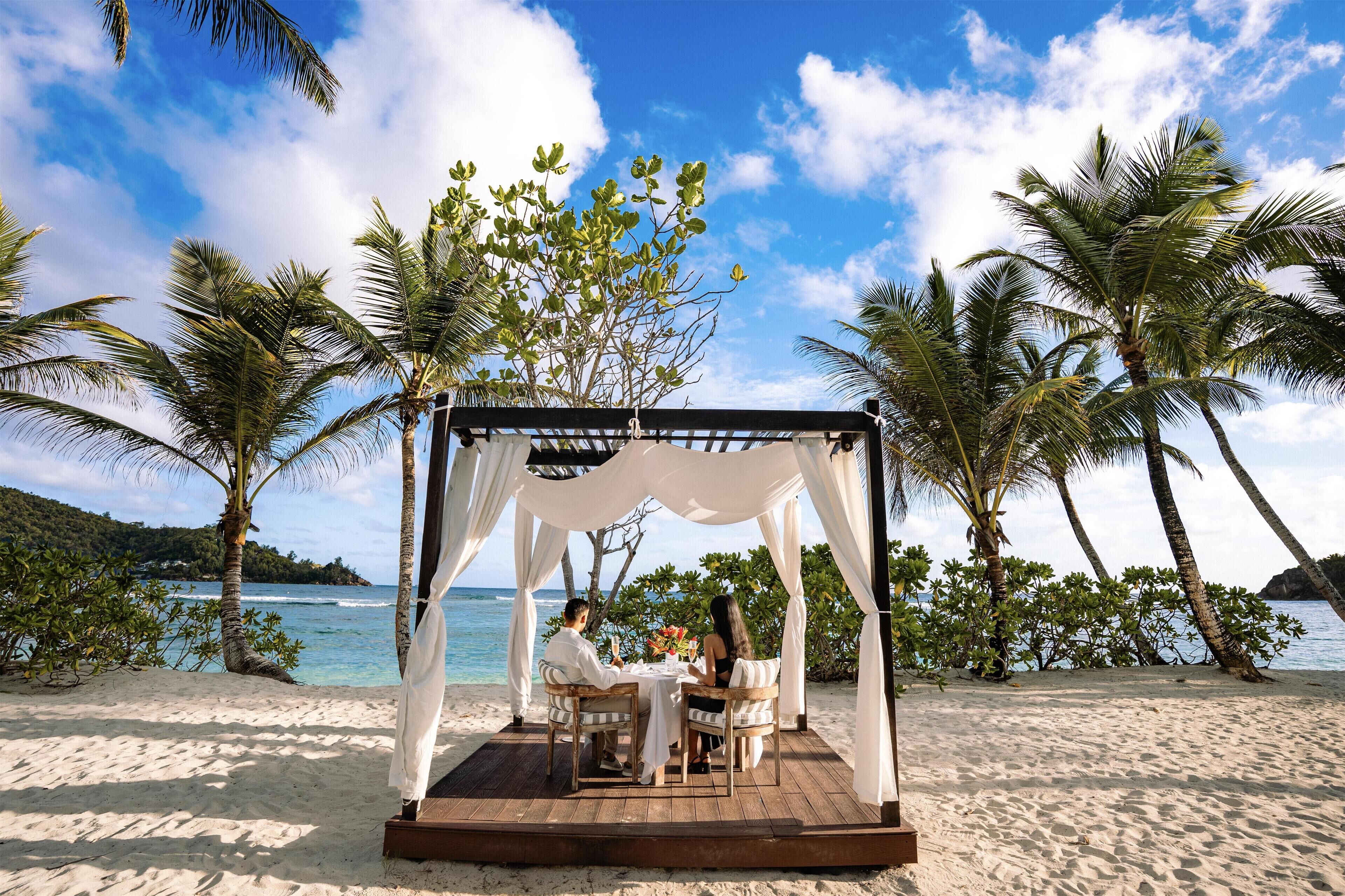 A beachside gazebo with a table and chairs, surrounded by palm trees and overlooking the ocean.
