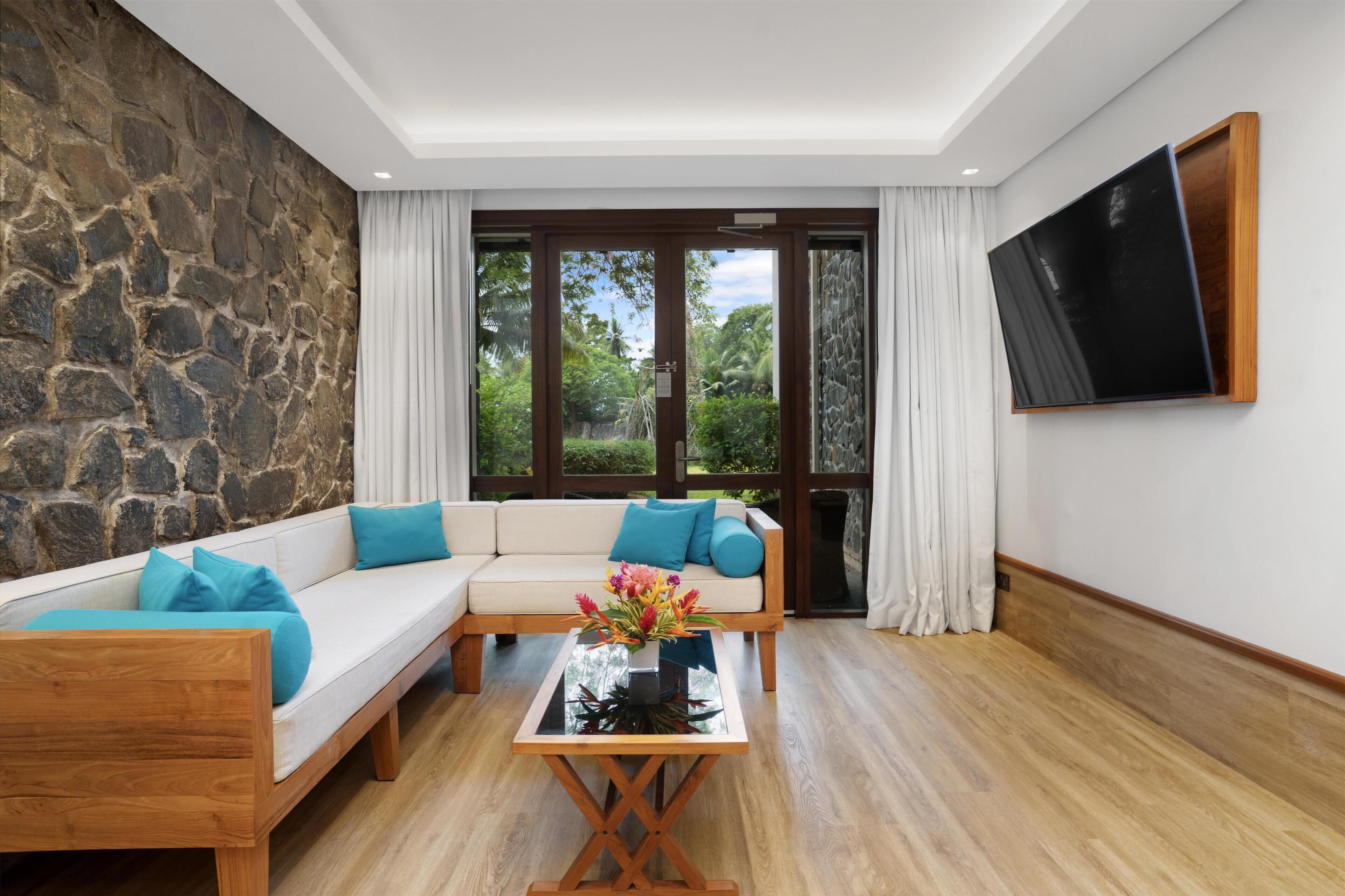 Ein modernes Wohnzimmer mit Steinwand, Holzboden, einem Ecksofa mit blauen Kissen, einem Glastisch mit Blumen und einem an der Wand montierten Fernseher.