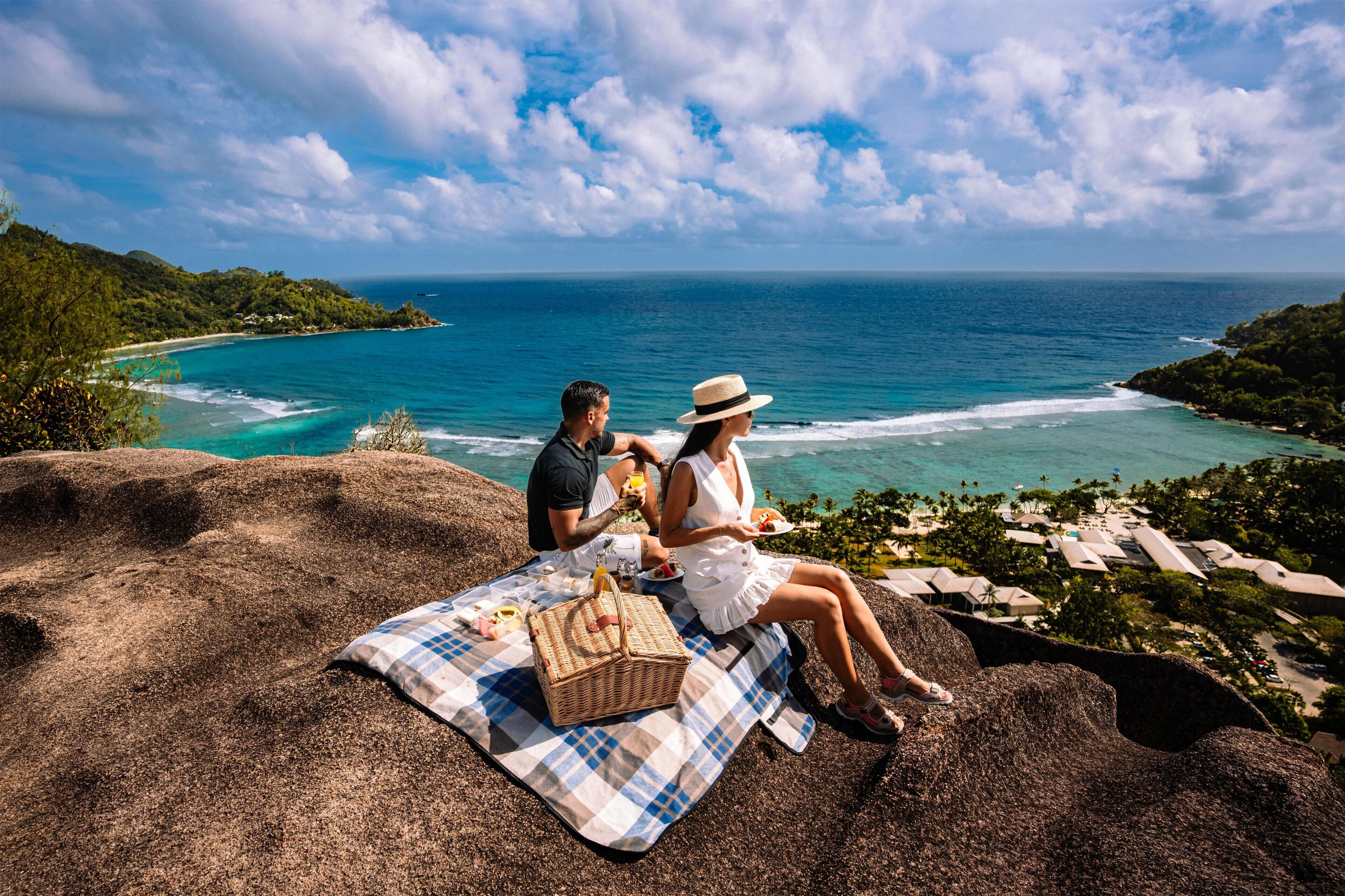 A couple on a blanket overlooking a beach with a clear view of the ocean and a resort in the background.