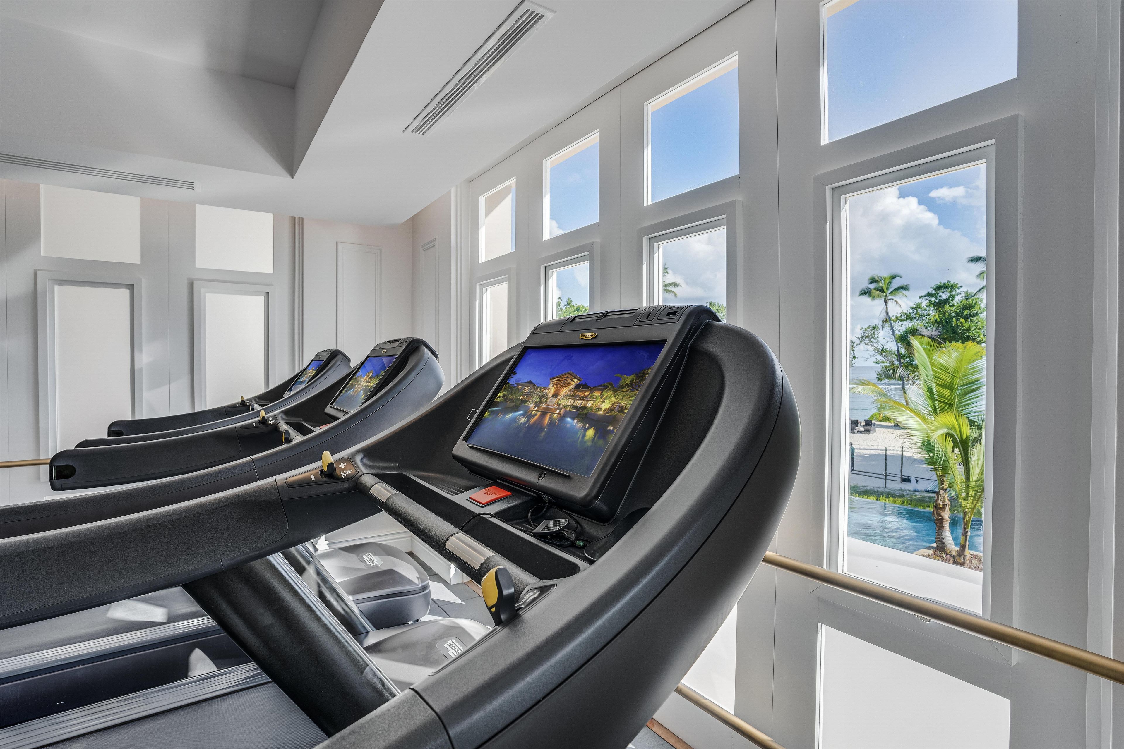 Un gimnasio con cintas de correr, amplias ventanas y vistas a un paisaje tropical.