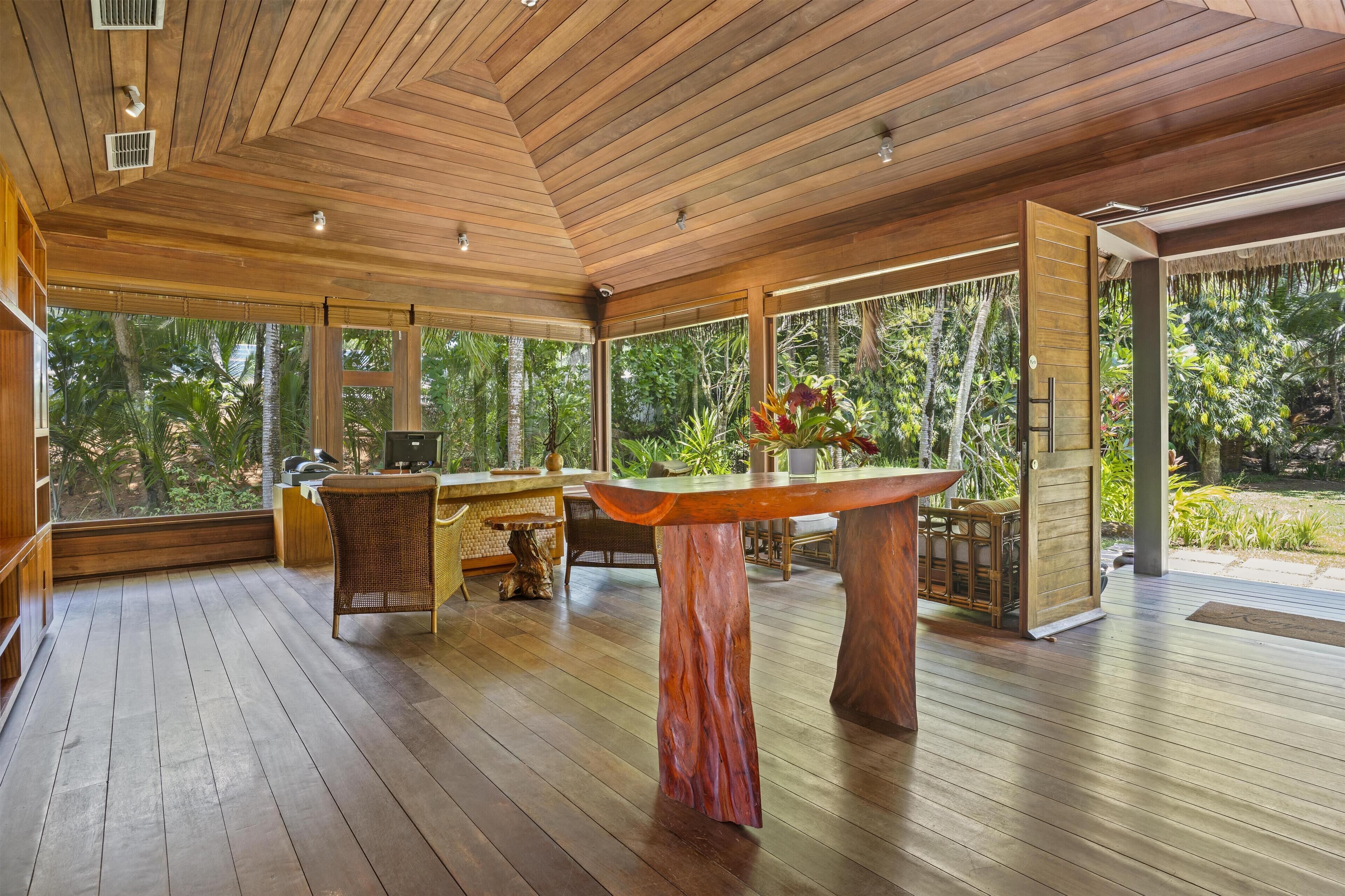 A spacious room with a wooden ceiling and floor, large windows offering a view of lush greenery outside.
