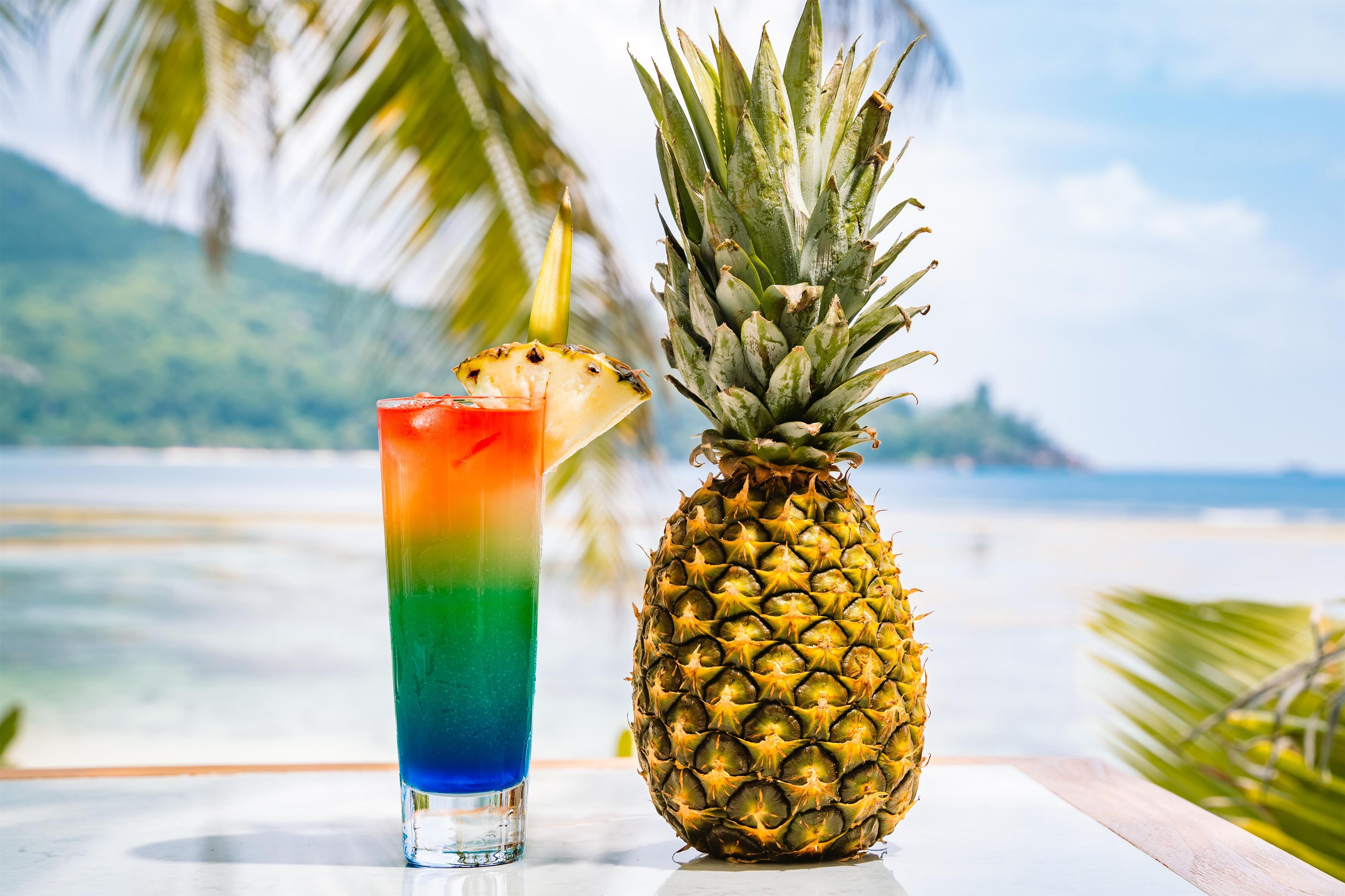 Un vaso de bebida tropical con una rodaja de piña en el borde, junto a una piña entera sobre una mesa, con una playa y palmeras al fondo.