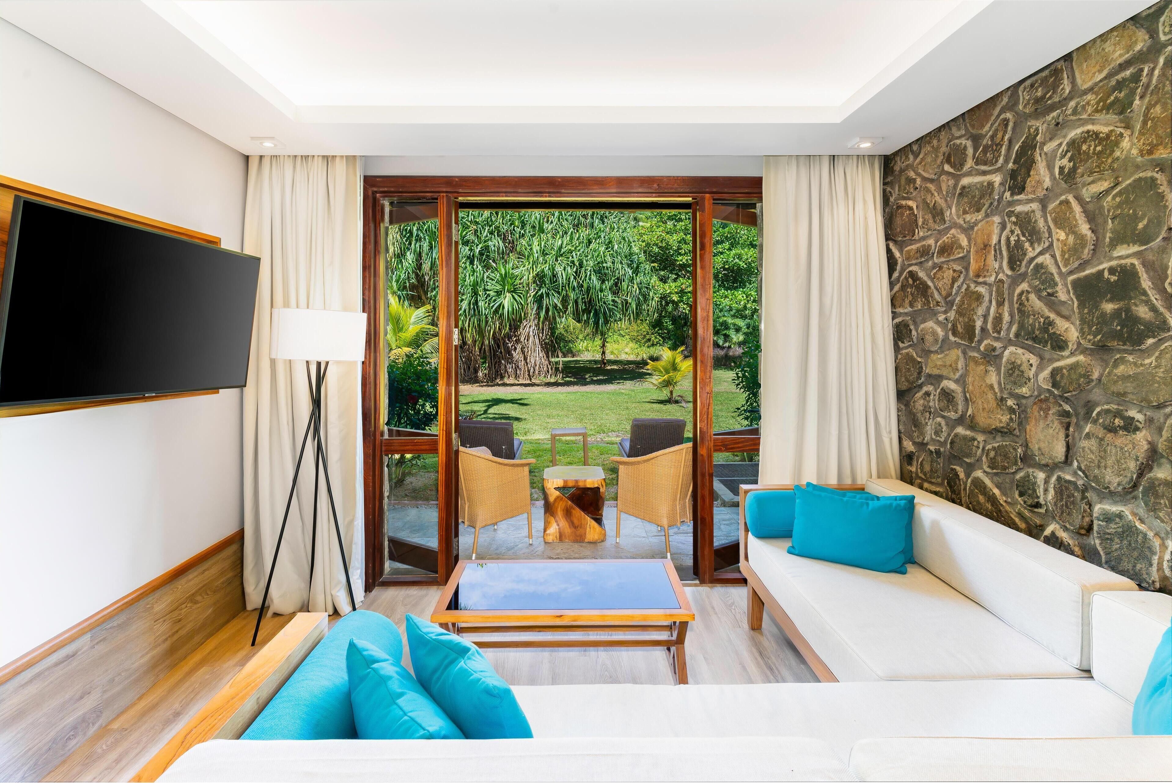 Une chambre d’hôtel moderne avec une grande télévision, un canapé avec des coussins bleus, une table basse en bois et un mur en pierre.