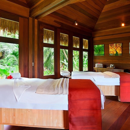 A room with two beds, large windows, wooden walls, and a view of greenery.