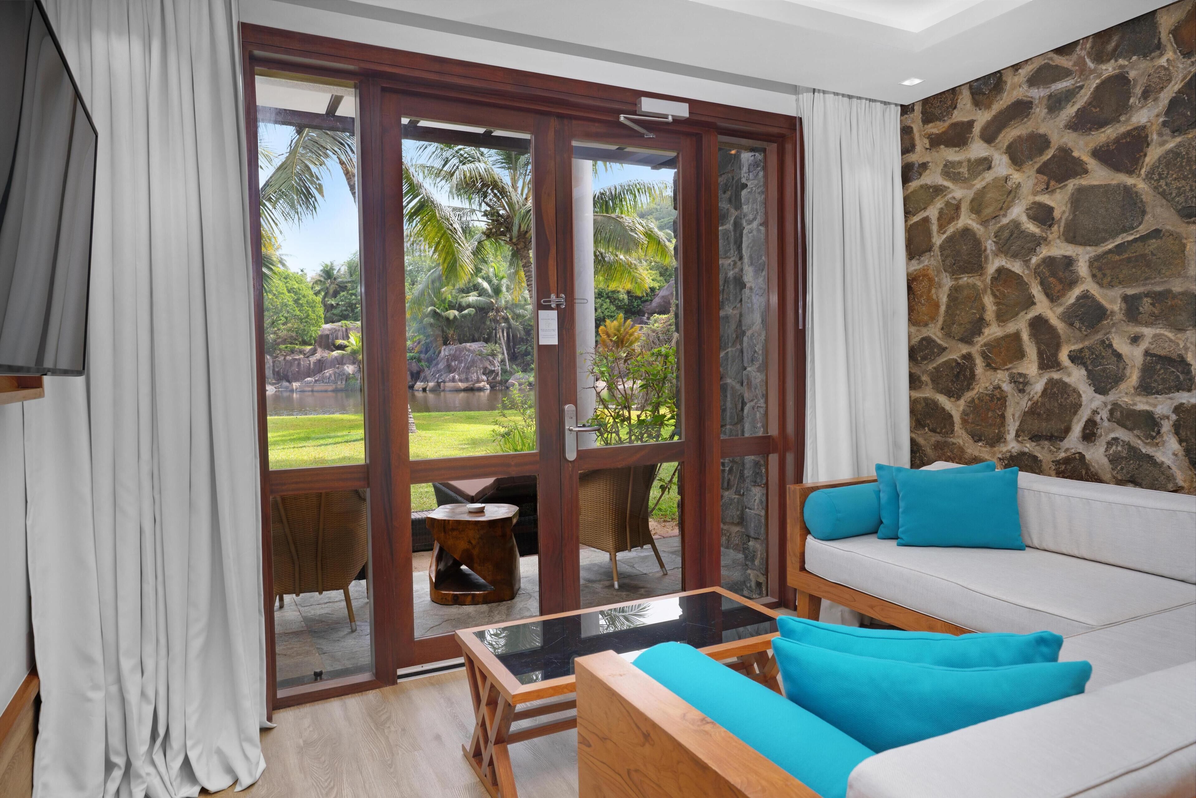 Un saló modern amb una porta de vidre que dona a un jardí tropical, un sofà blanc amb coixins blaus i una taula de centre de fusta.