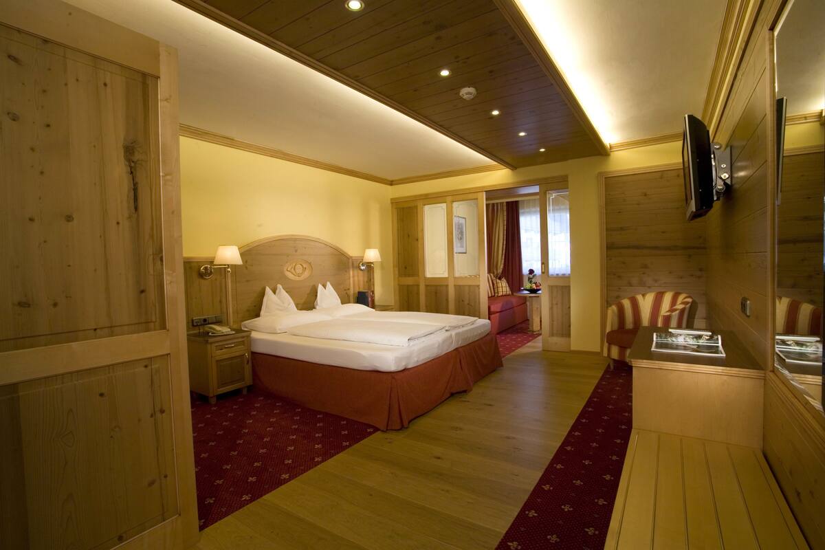 A hotel room with a wooden floor, a large bed with white linens, a wooden headboard, a bedside table with a lamp, a small desk with a mirror, and a television mounted on the wall.