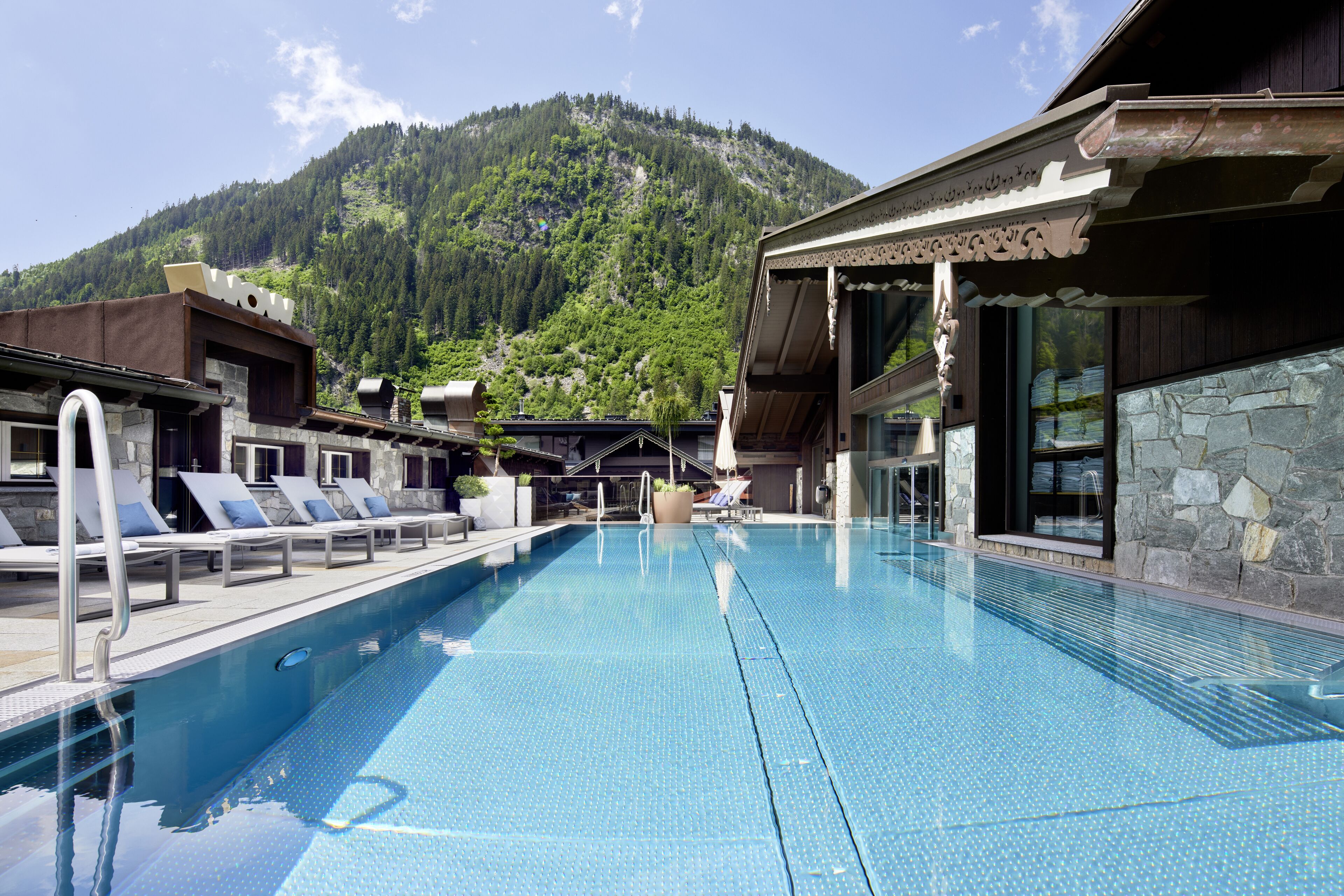 A swimming pool with lounge chairs, a stone wall, and a mountain backdrop.
