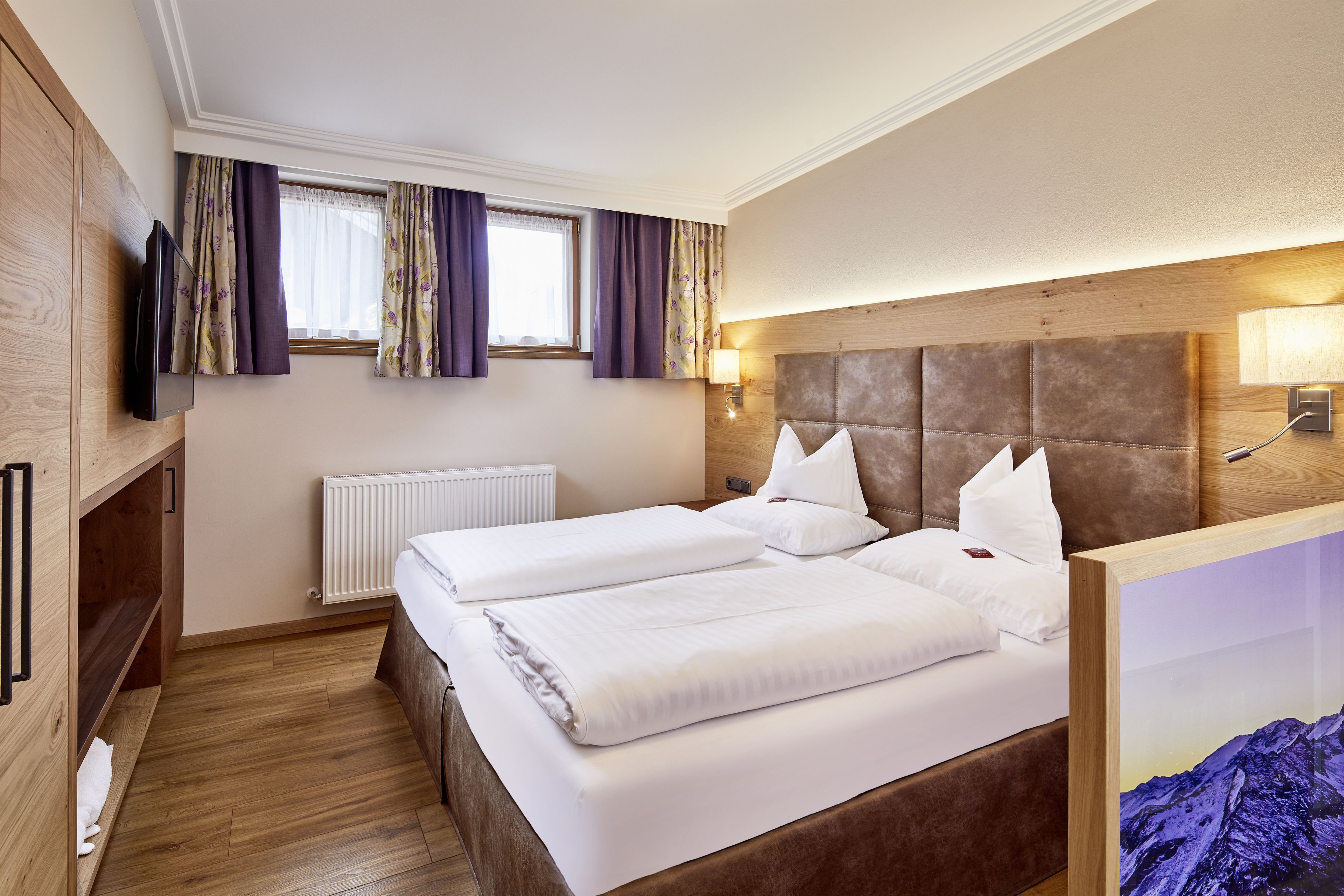 A hotel room with two beds, wooden flooring, a TV, a window with curtains, a radiator, and a headboard with a light fixture.