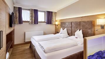 A hotel room with two beds, wooden flooring, a TV, a window with curtains, a radiator, and a headboard with a light fixture.