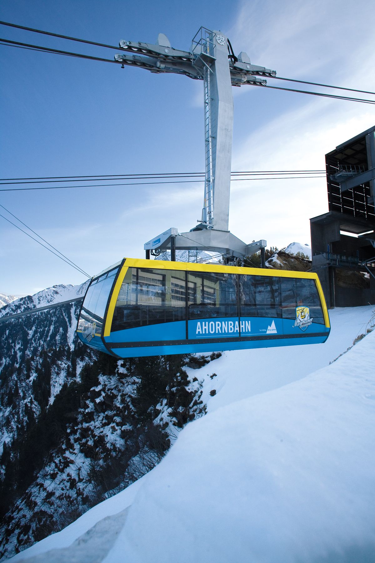 A cable car with "AHORNBAHN" written on it, suspended by cables against a snowy mountain backdrop.