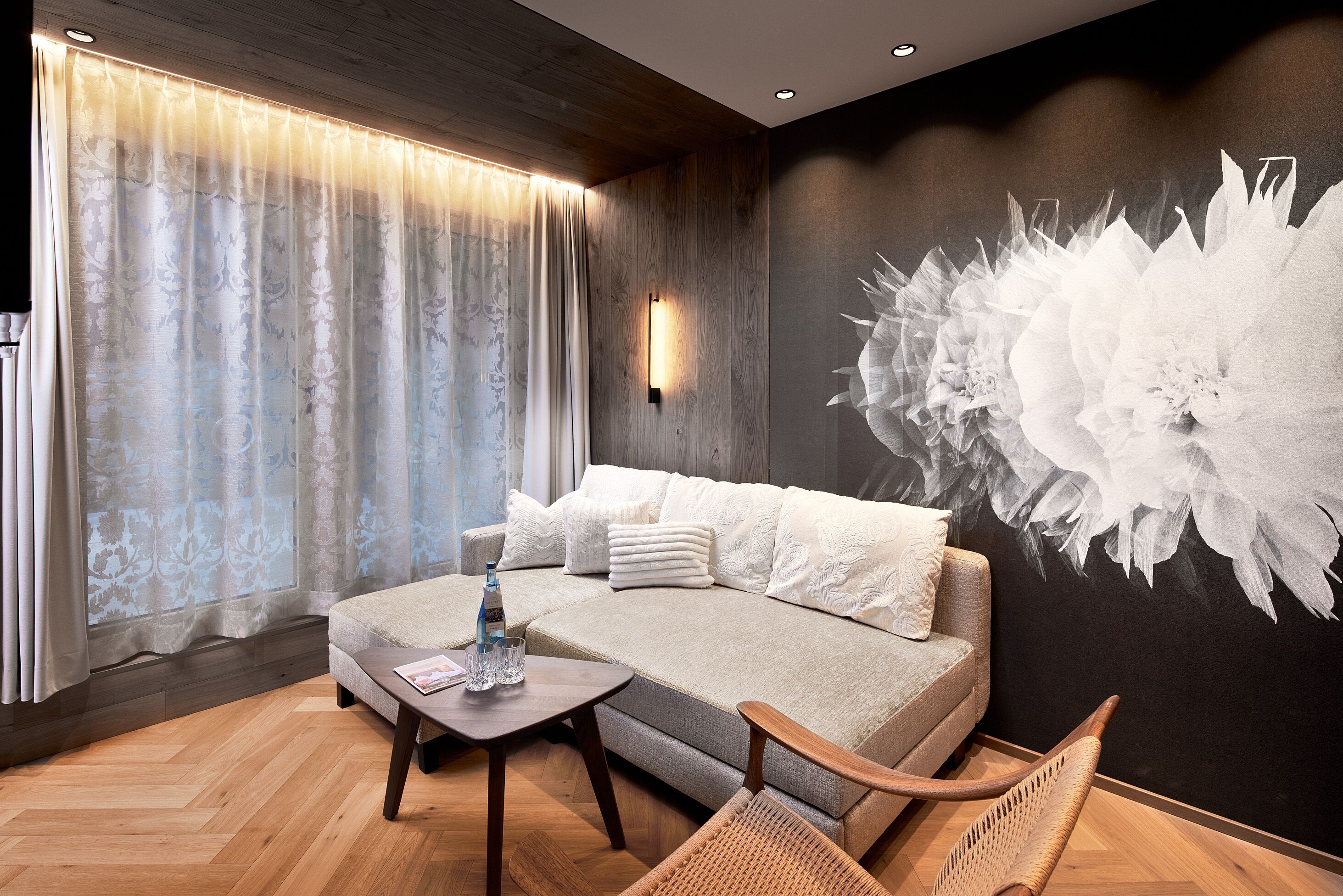 Een moderne kamer met een bank, een salontafel en een stoel. Er is een groot raam met gordijnen, een wandlamp en een groot bloemenkunstwerk.