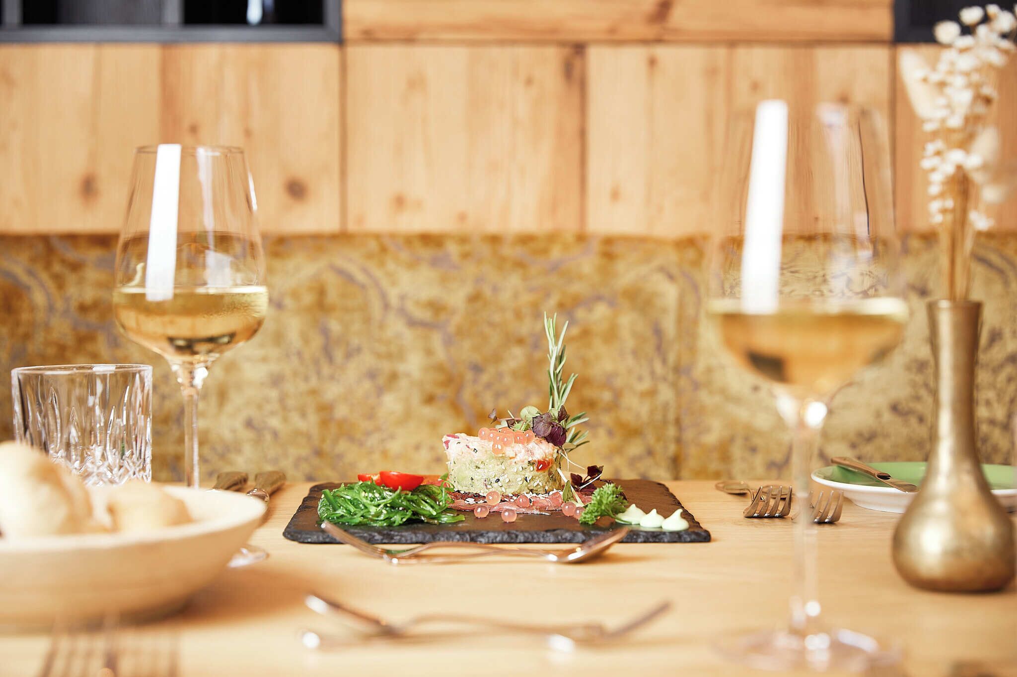 Une table à manger avec une assiette de nourriture, deux verres de vin blanc et un vase rempli de fleurs.
