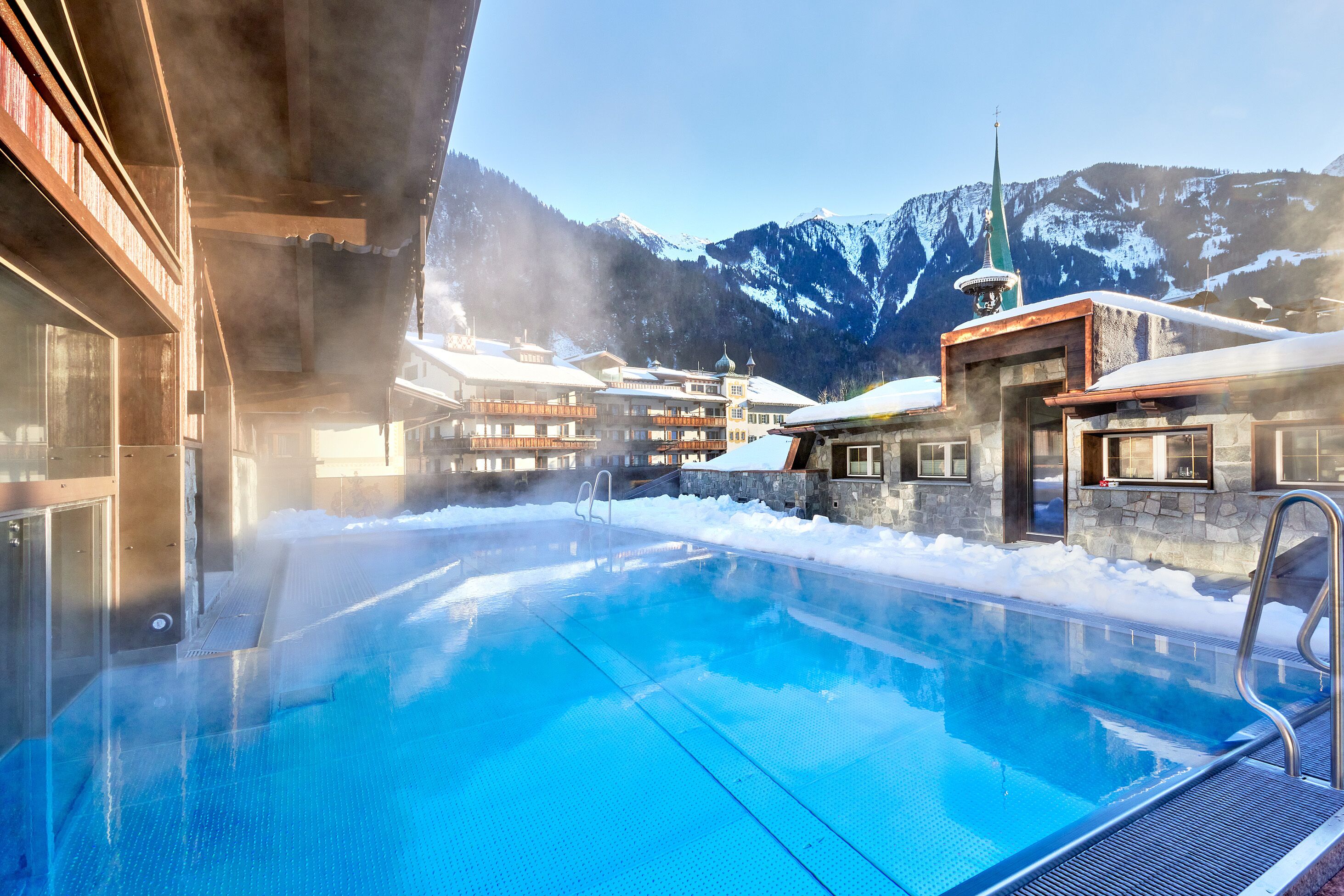 Un espace piscine avec une eau d’un bleu limpide, entouré de bâtiments aux façades en pierre et aux toits enneigés, le tout se détachant sur un paysage de montagnes aux sommets couverts de neige.