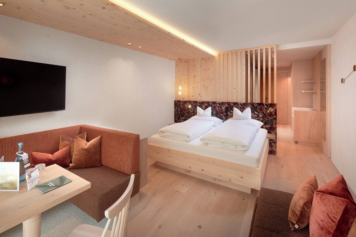A modern hotel room with a wooden ceiling, a bed with white linens, a seating area with a table, and a flat-screen TV mounted on the wall.