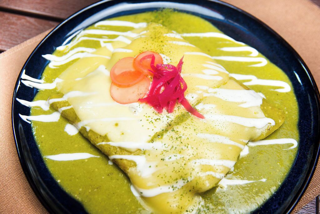 Une assiette d’enchiladas nappées de sauce verte et de sauce blanche, garnie de tomates et d’oignons rouges.