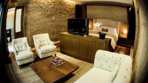 A cozy hotel room with a stone wall, a bed with white linens, a television on a wooden cabinet, and two white armchairs. - Casa Madero Hotel Boutique (Morelia)
