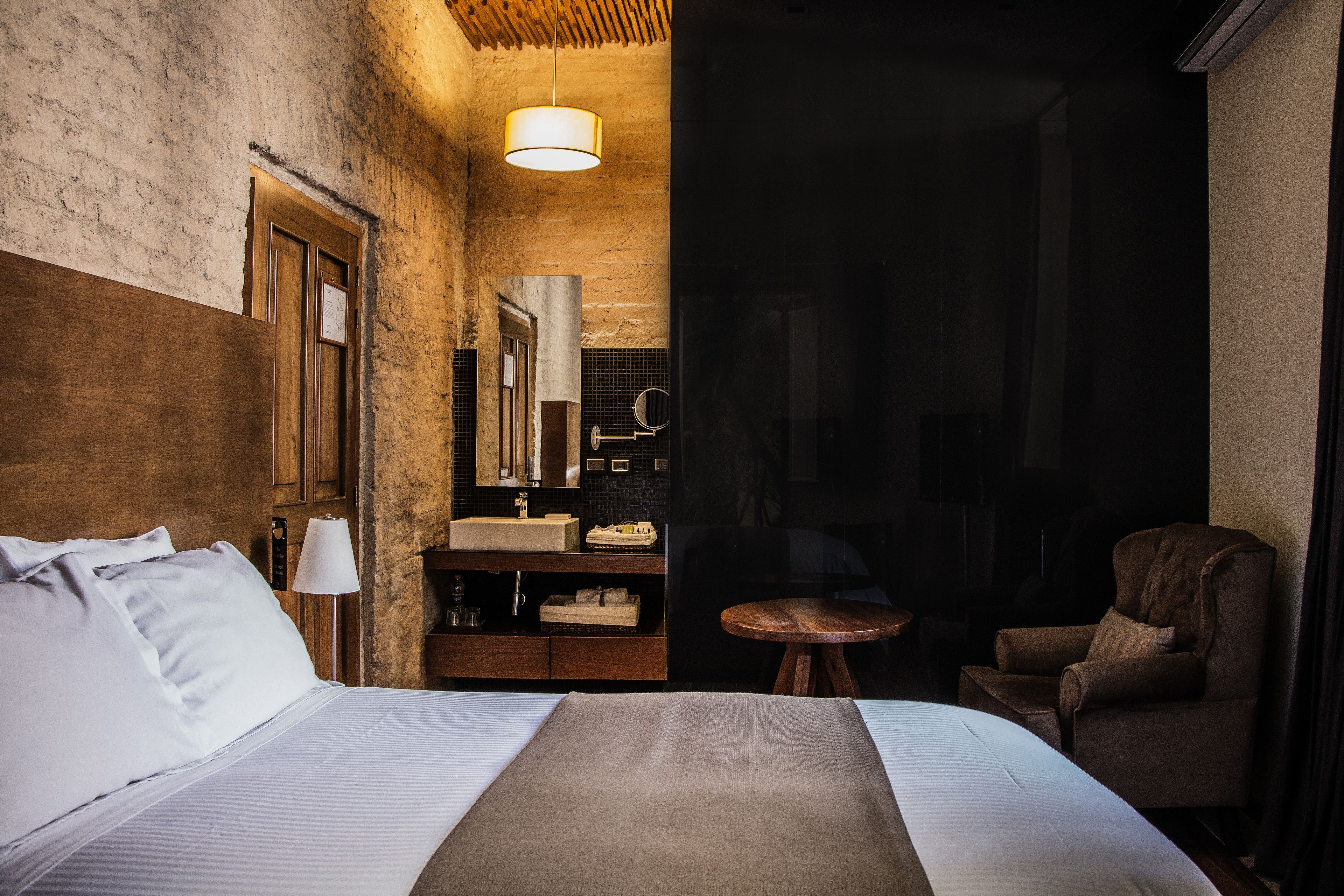 Habitación de hotel con una cama grande, cabecera de madera, una mesita redonda pequeña, una silla y un baño con espejo y lavabo.