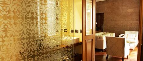 A modern bathroom with a glass shower enclosure and a wooden door leading to a bedroom with a bed and a chair.