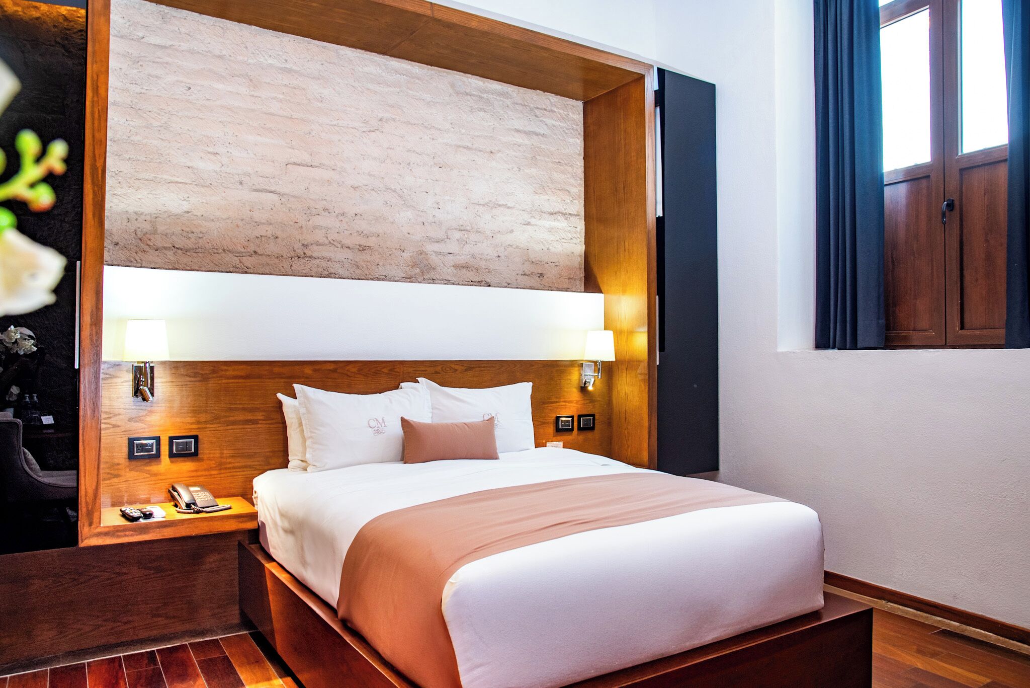 A hotel room with a wooden headboard, a large bed with white and pink bedding, a bedside table with a lamp, and a nightstand with a phone and remote control.