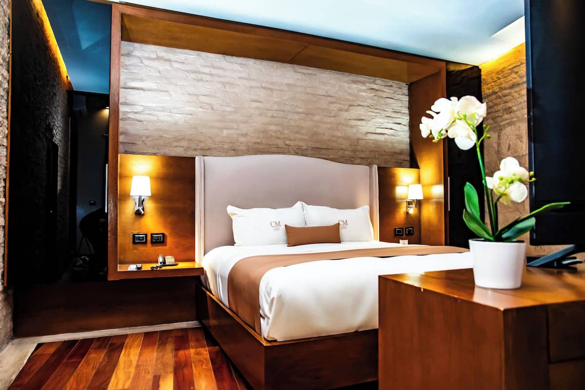 A modern hotel room with a wooden bed, a nightstand, a desk, and a potted plant.