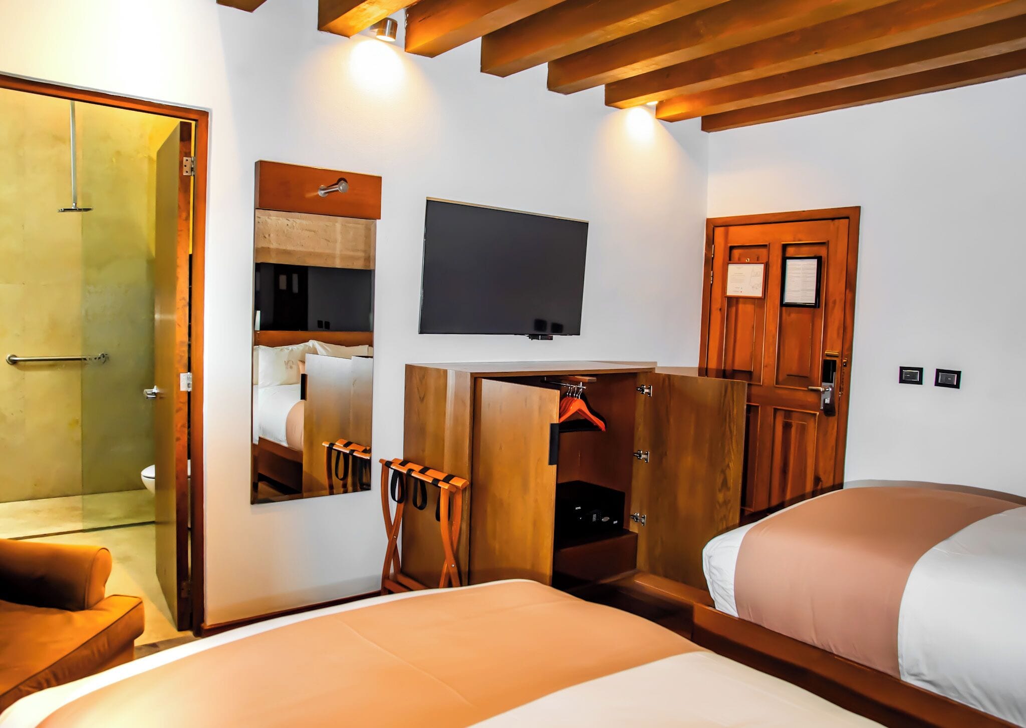 A hotel room with a bed, a TV mounted on the wall, a wooden wardrobe, a mirror, and a shower area.