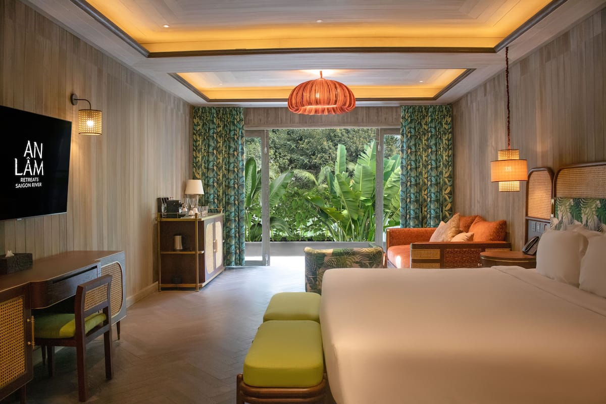 A modern hotel room with a large bed, a desk, a television, and a view of lush greenery.