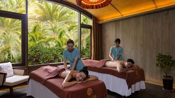 A spa room with two massage tables, a large window with a view of tropical plants, and a wooden ceiling.
