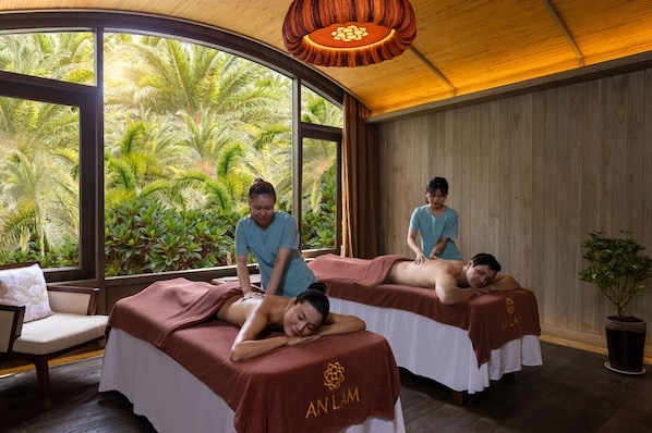 A spa room with two massage tables, a large window with a view of tropical plants, and a wooden ceiling.