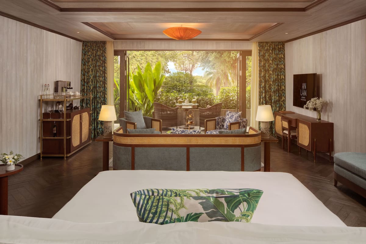 A hotel room with a large bed, a sitting area with chairs, a small table, and a view of greenery outside.