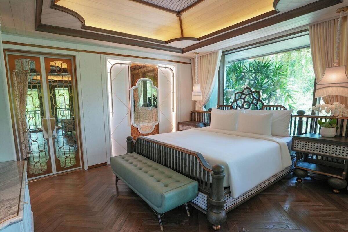 A spacious bedroom with a large bed, a bench, a nightstand, and a view of greenery.