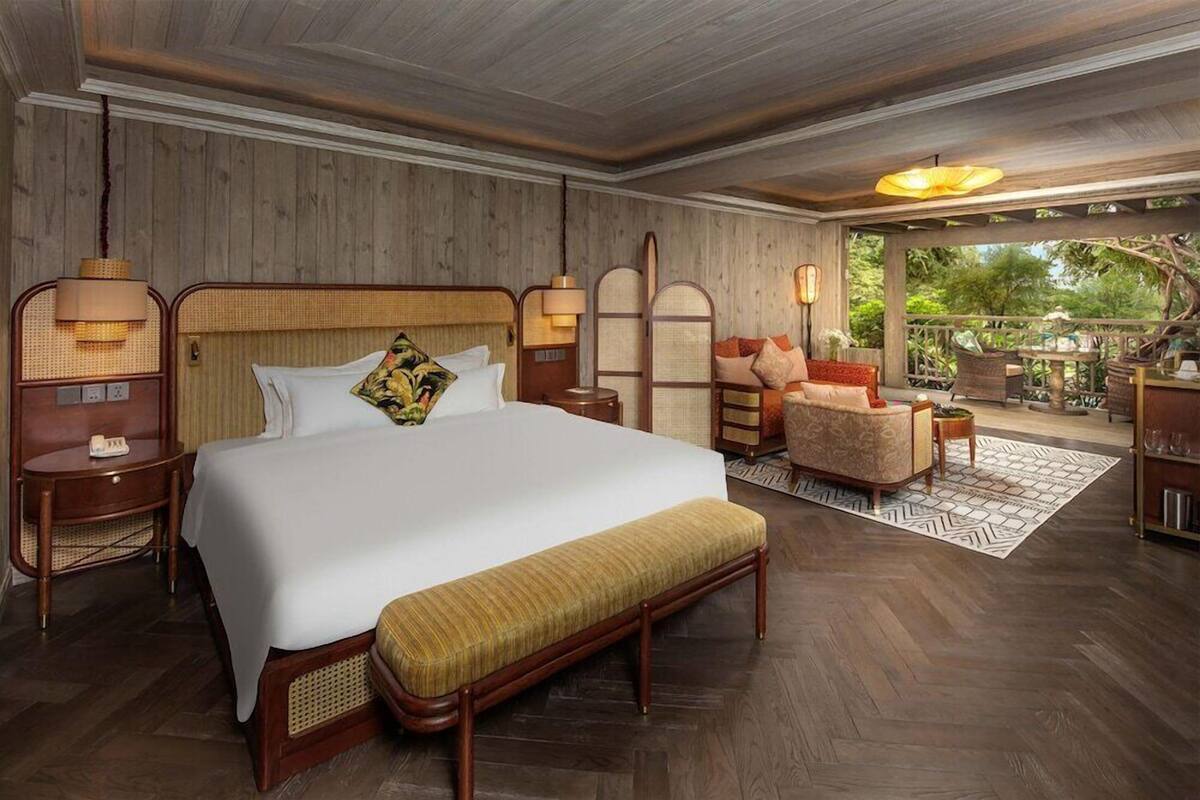 A spacious bedroom with a large bed, a wooden headboard, bedside lamps, a bench, and a view of the outdoors.