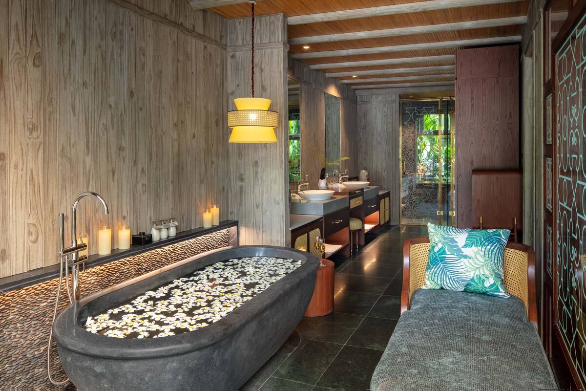 Ein modernes Badezimmer mit einer großen Badewanne, gefüllt mit weißen Blumen, einem hölzernen Waschtisch und einer Sitzecke mit Kissen.