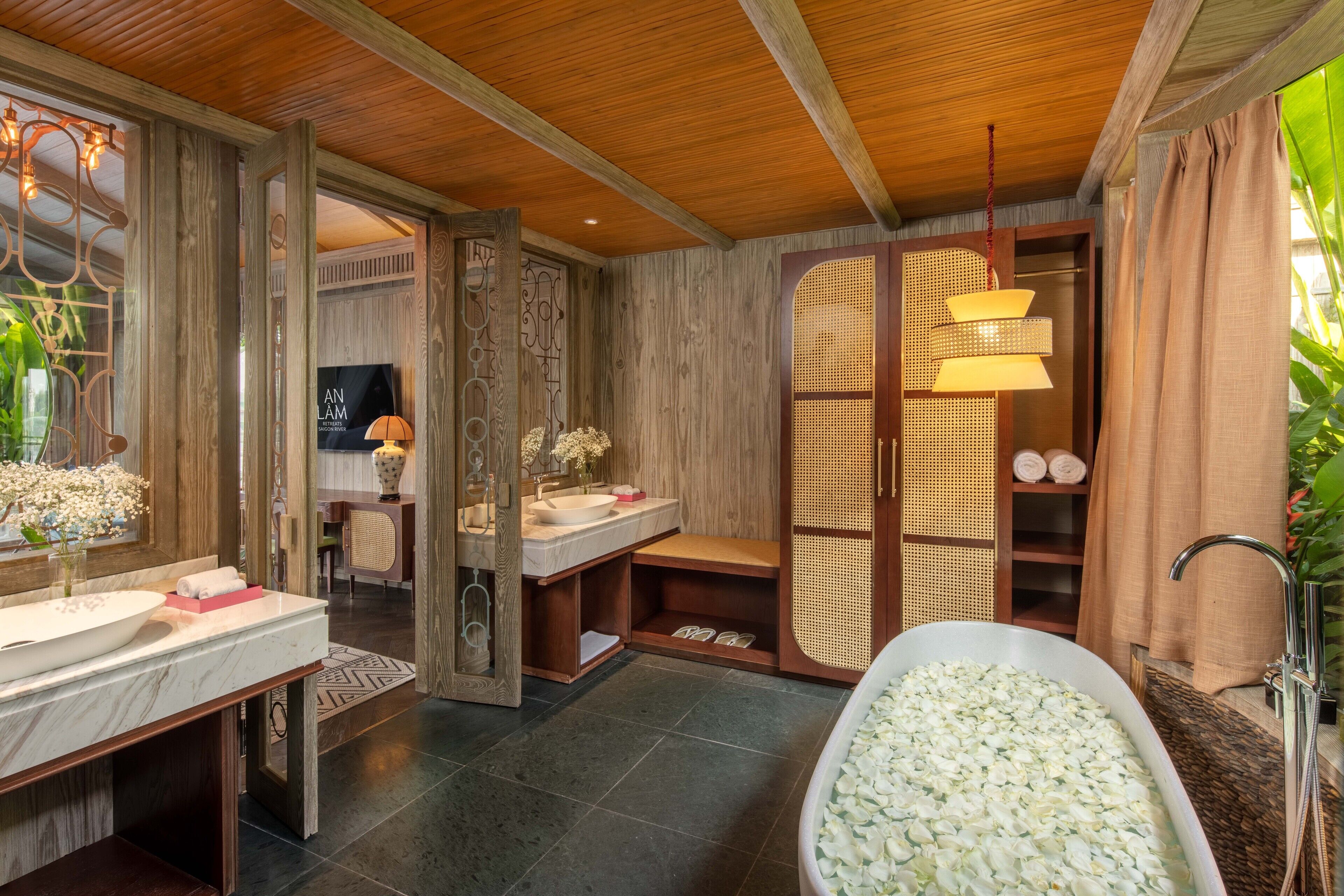 A bathroom with a wooden ceiling, a large bathtub filled with white flowers, a wooden vanity with a mirror, and a wicker shelf.