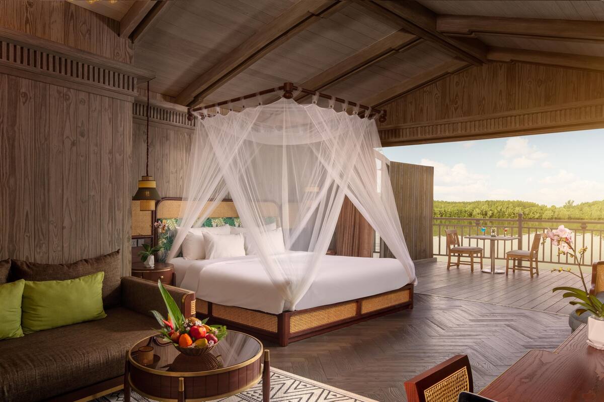 A bedroom with a four-poster bed, a sitting area with a sofa, a dining table with chairs, and a view of a lake.