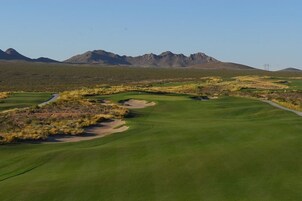 Un campo de golf con cielo despejado, fairways verdes, búnkeres de arena y montañas al fondo.