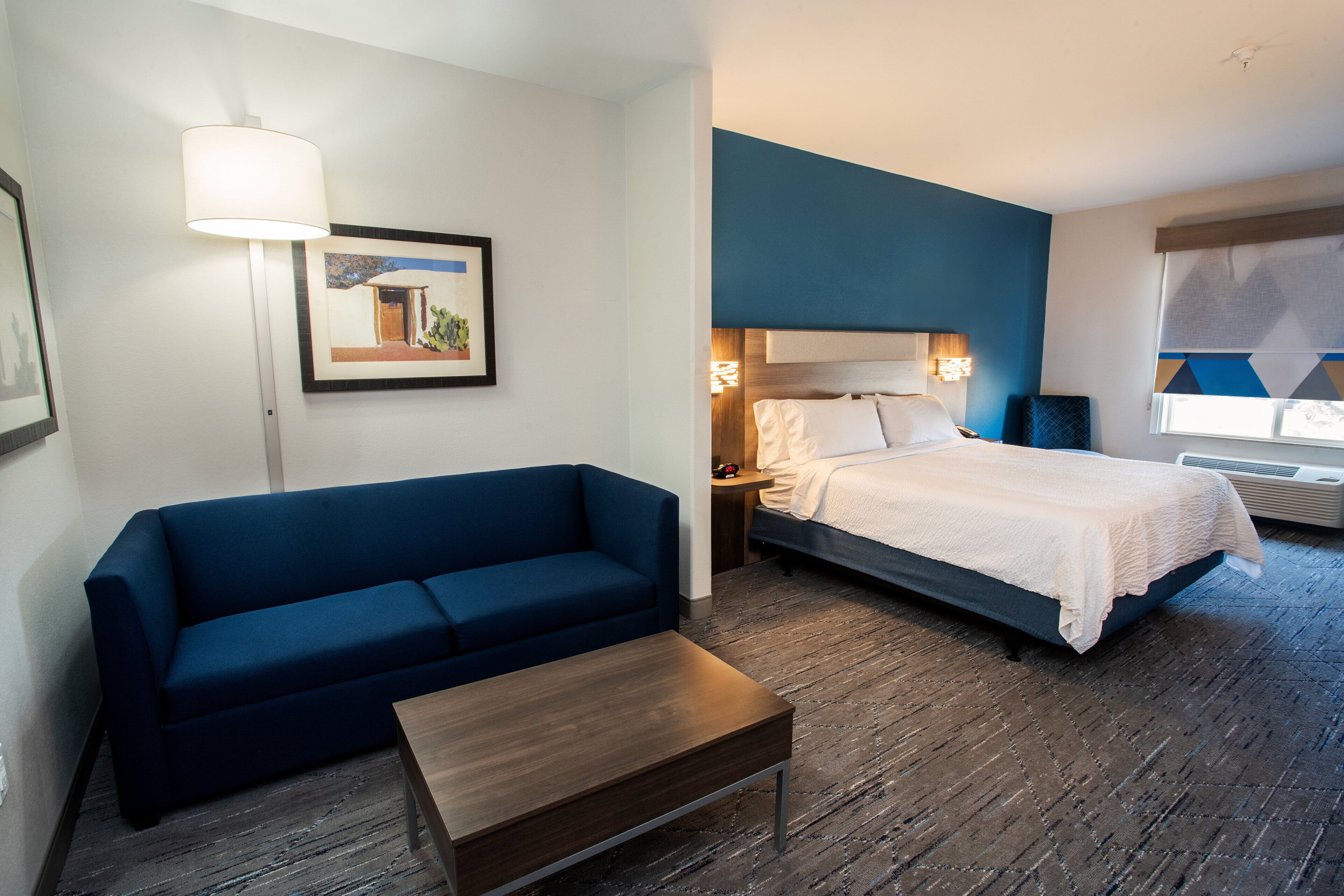 A hotel room with a blue sofa, a wooden coffee table, a bed with white bedding, a nightstand with a lamp, and a framed picture on the wall.