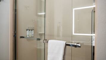 A glass-enclosed shower with a towel hanging on a metal bar, a glass shelf with toiletries, and a mirror reflecting the shower area.