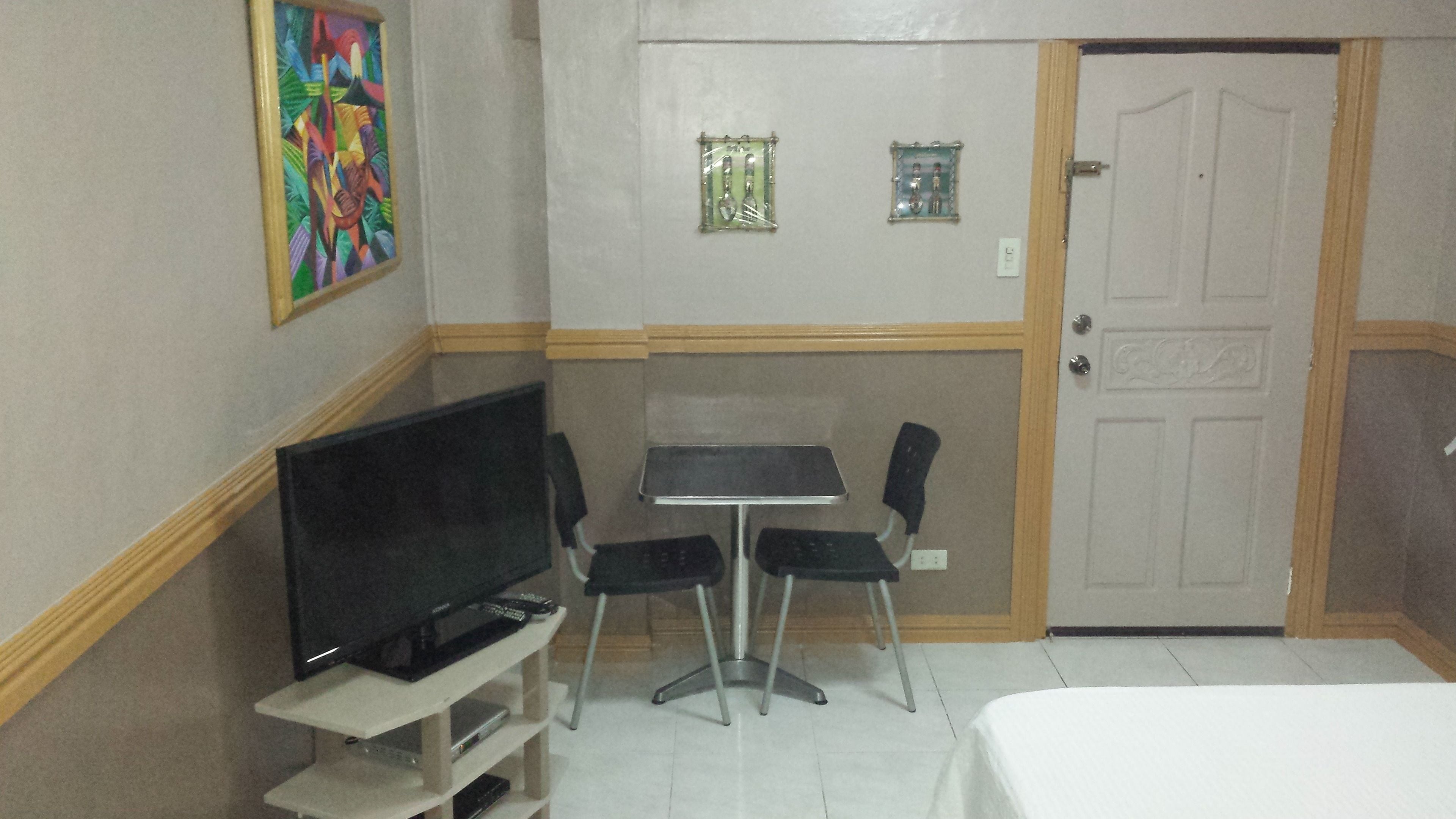 A room with a television on a stand, a small table with two chairs, a door, and a wall-mounted light fixture.