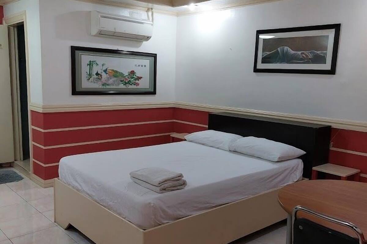 Ein Hotelzimmer mit einem Bett, einem Schreibtisch und einem Stuhl. Es gibt einen Deckenventilator, eine Klimaanlage und zwei gerahmte Bilder an der Wand.