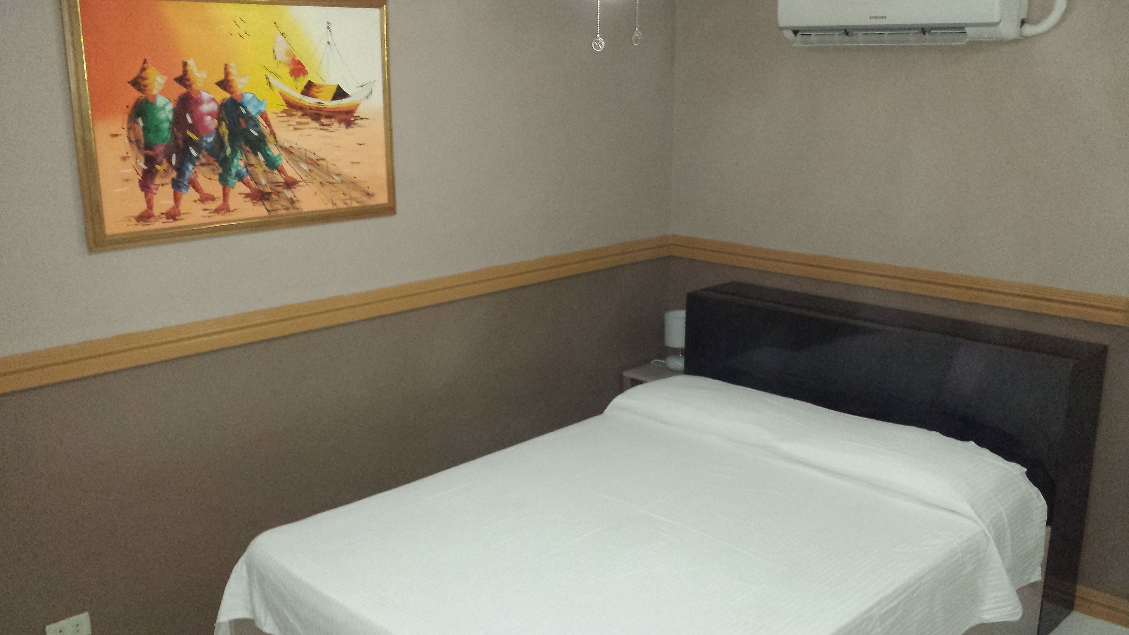 A hotel room with a single bed, a painting on the wall, and an air conditioning unit above the bed.