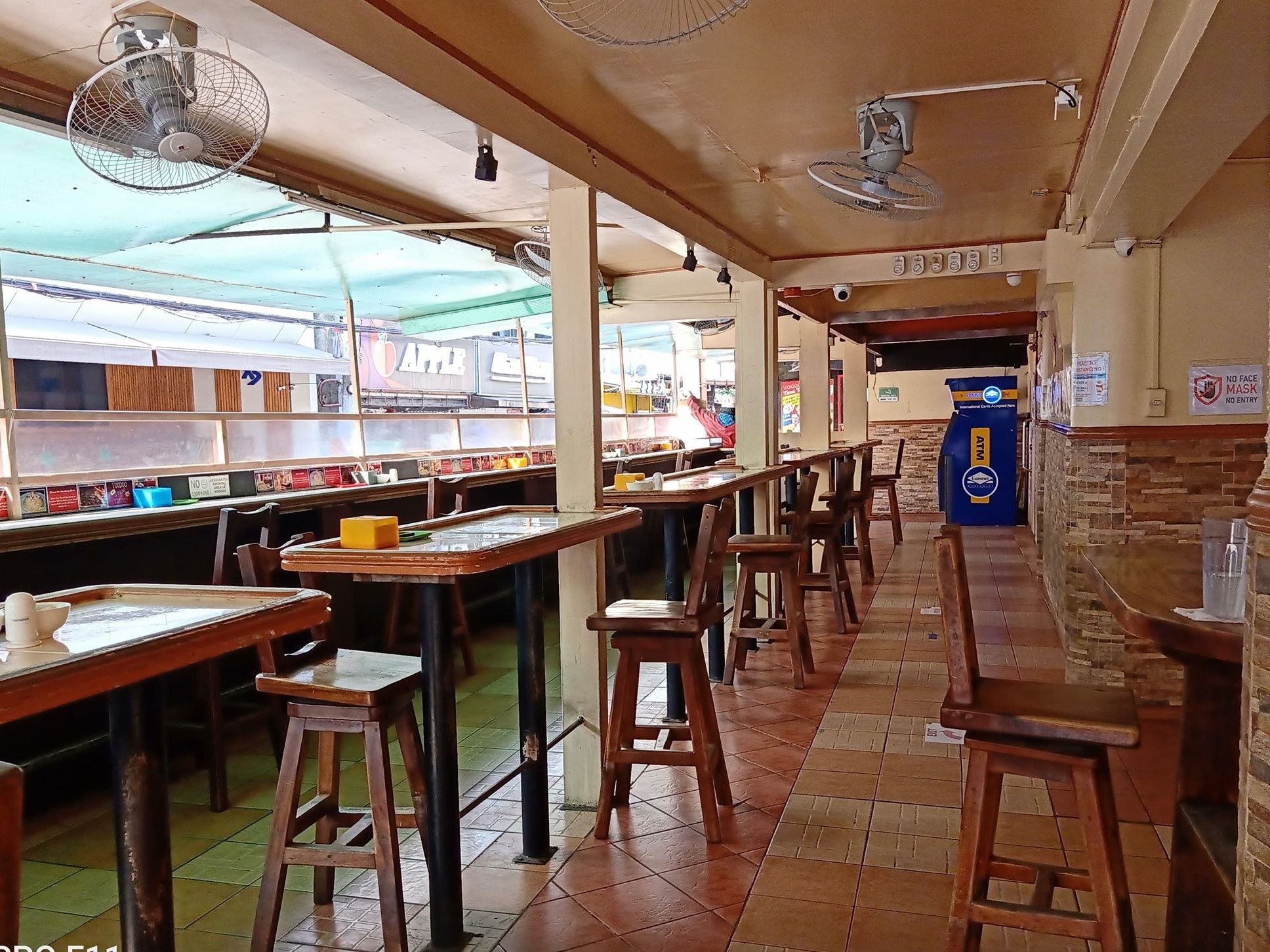 A restaurant with long communal tables, bar stools, and a counter with an ATM.