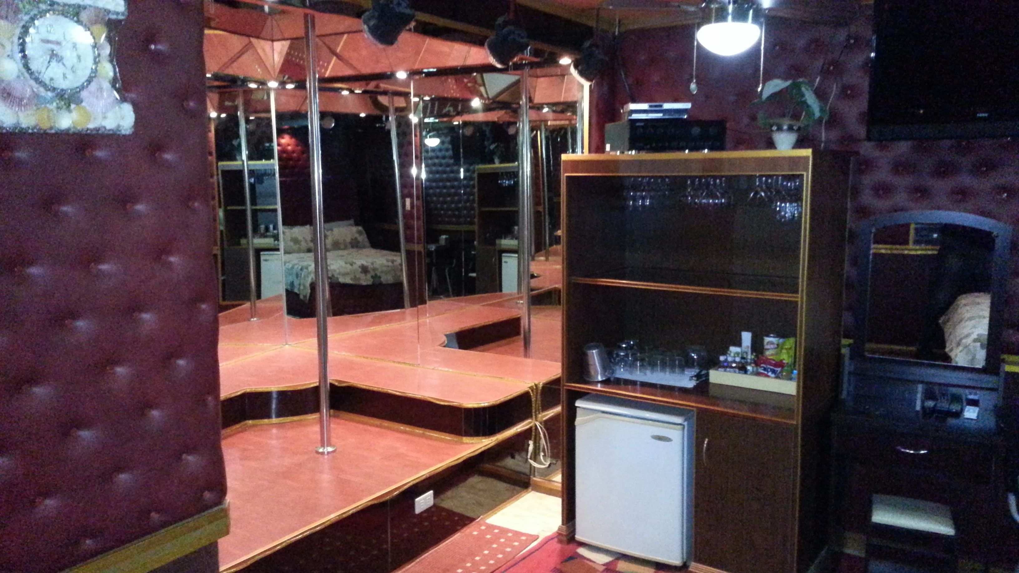 A room with a mirrored wall, a small refrigerator, and a shelf with various items.