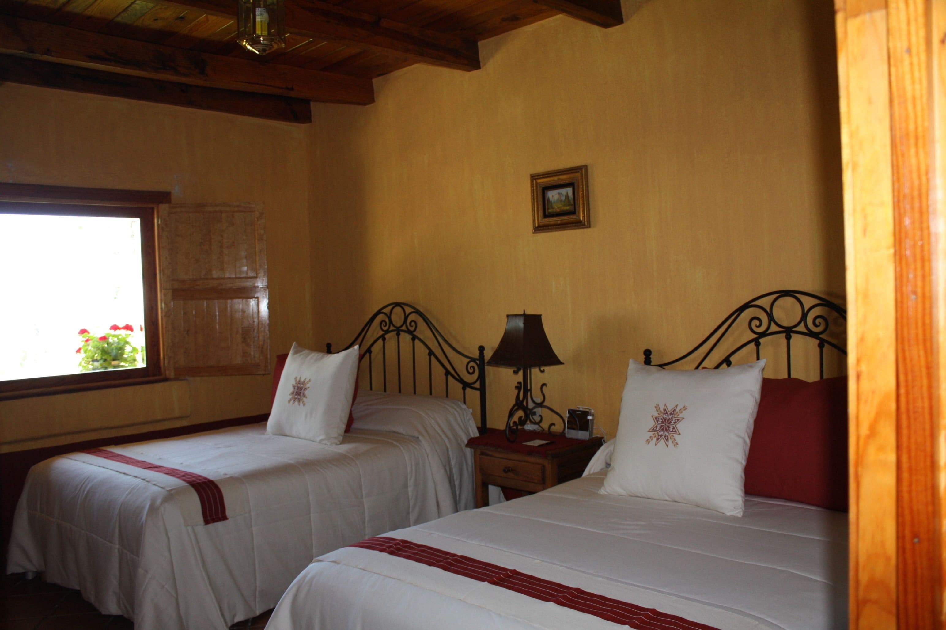 A hotel room with two beds, wooden beams on the ceiling, a window with a view of flowers, and a small table with a lamp.