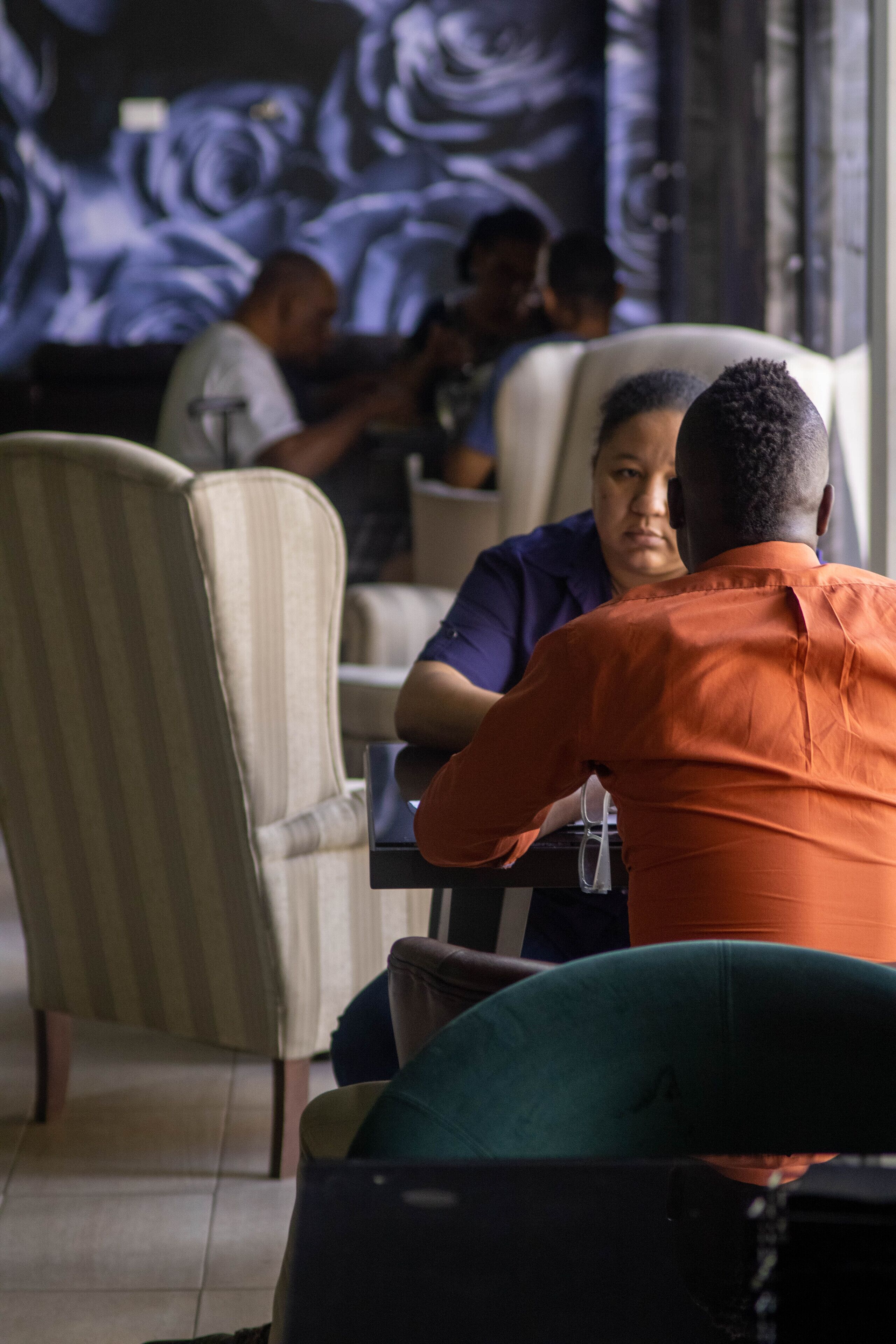 Una persona con camisa naranja sentada en una mesa con una computadora portátil, en una habitación con sillas a rayas y una pantalla grande mostrando arte abstracto.