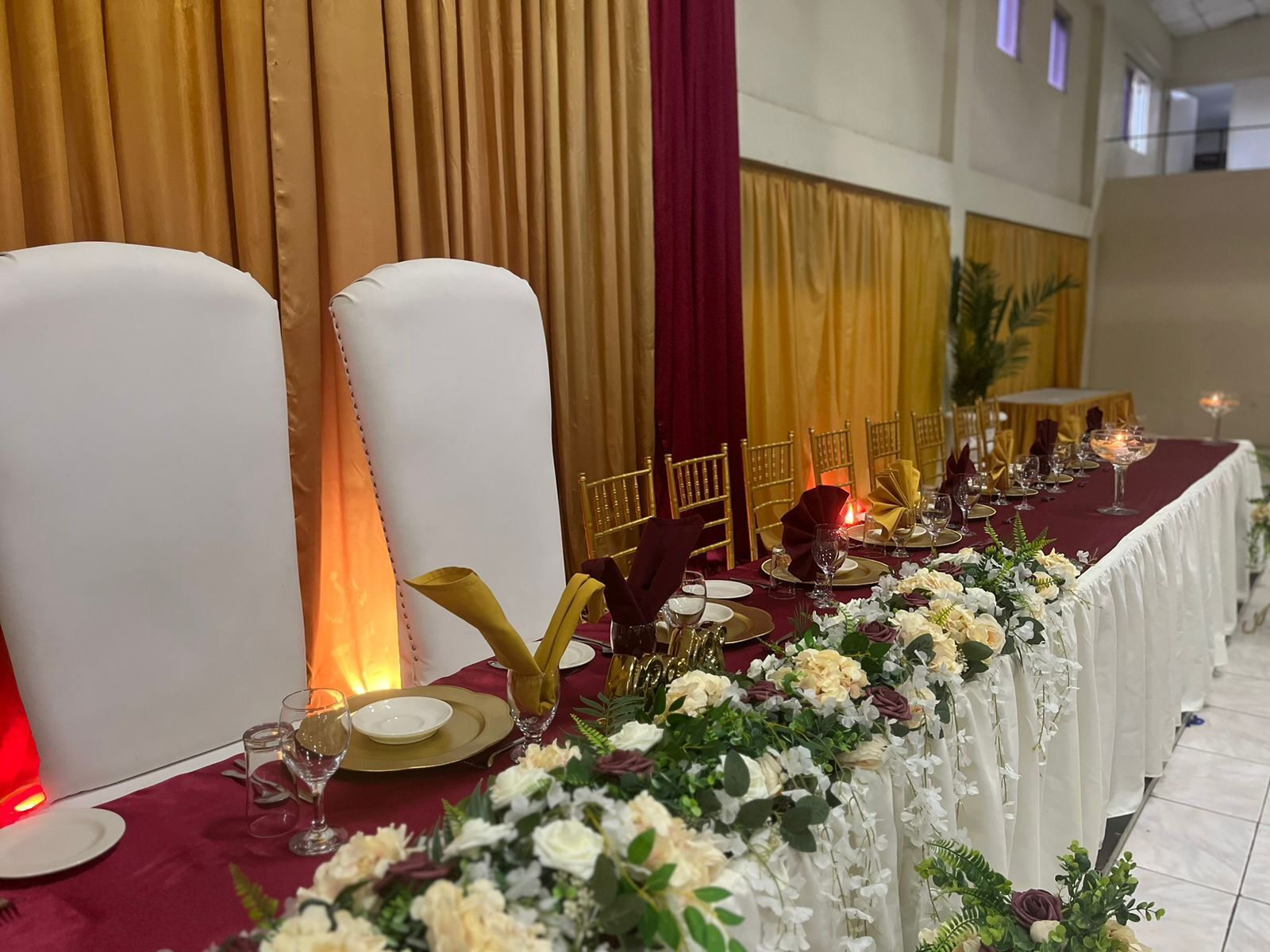 A banquet table set with white chairs, gold cutlery, and floral centerpieces.