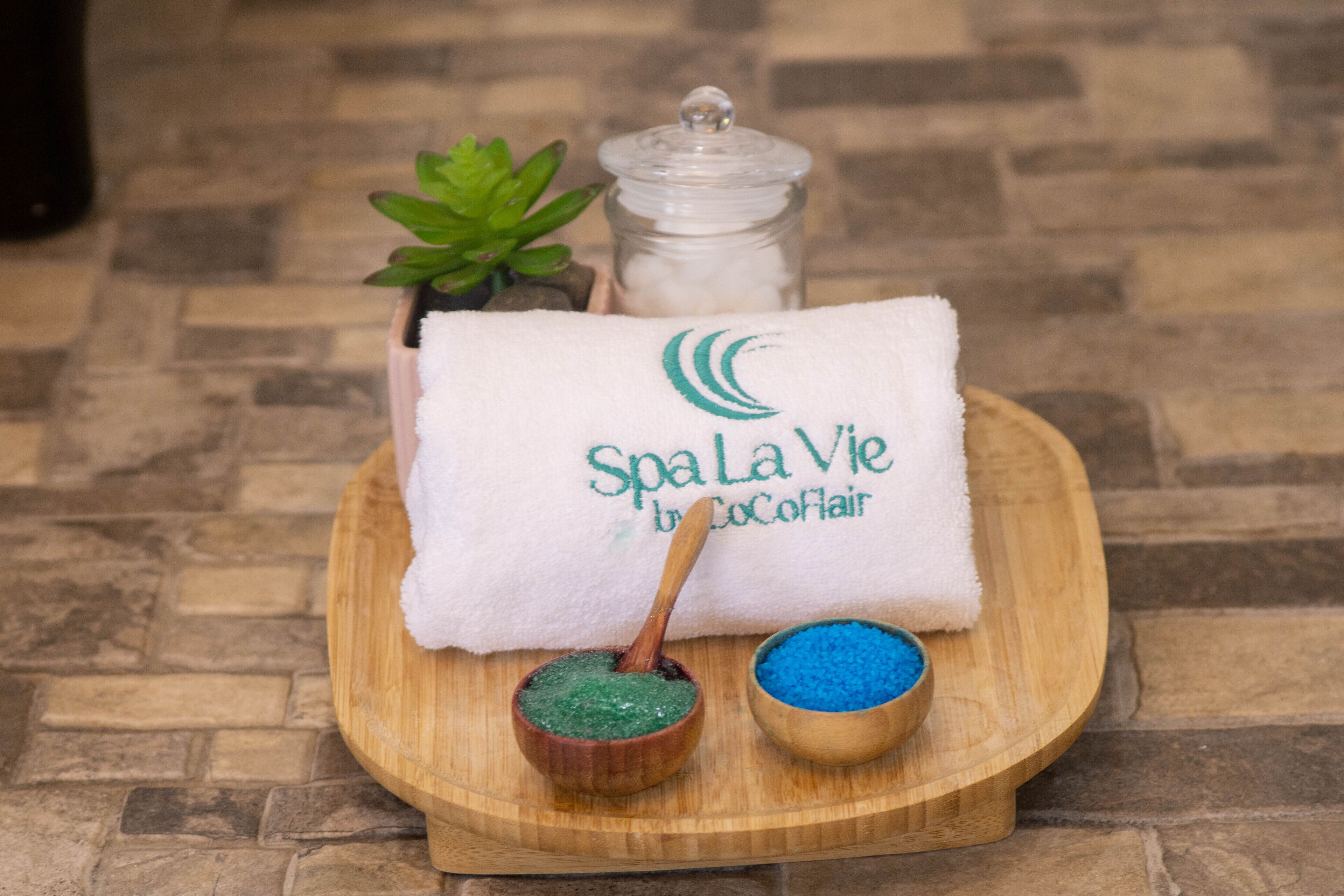 Toalla de spa con el logo Spa La Vie by Cocofair, una planta pequeña en maceta y dos cuencos de madera con sales de baño verdes y azules sobre una bandeja de madera.
