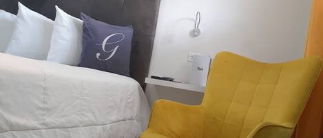 Un dormitorio con cama, cabecera, mesita de noche, una silla y una lámpara fijada a la pared.