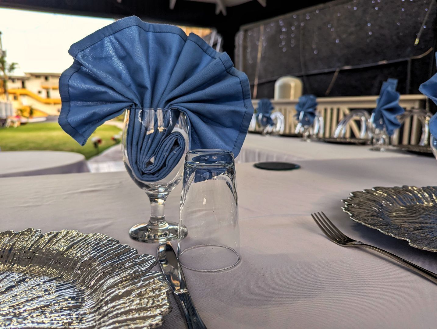 Una mesa dispuesta con una servilleta azul doblada, una copa de cristal para vino, una copa de cristal para agua, un plato de plata y un tenedor.